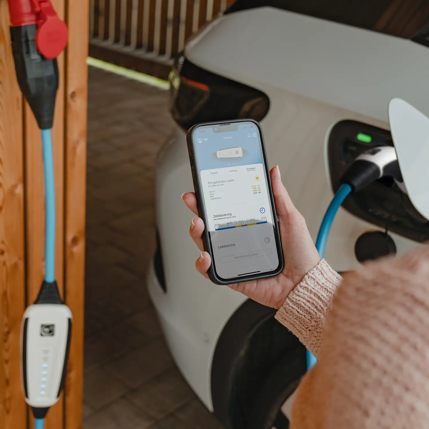 Elektroauto lädt mit NRGkick-Wallbox, Frau demonstriert App auf Smartphone.